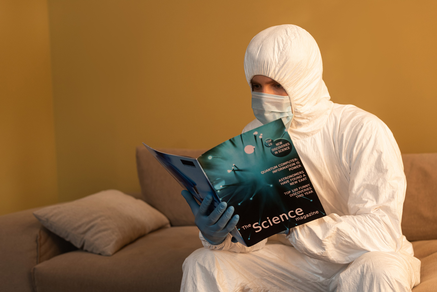 Man in hazmat suit, medical mask and latex gloves reading science magazine on sofa